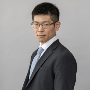 image of Ohyun wearing glasses standing in a grey suit, white dress shirt and  light blue tie