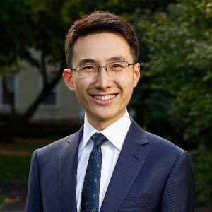 head shot of Xiang wearing glasses, a white collar shirt, blue pattern tie and blue blazer