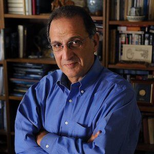 image of Dr Zogby standing in fron of a large bookcase wearing glasses and a blue dress shirt with his arms crossed 