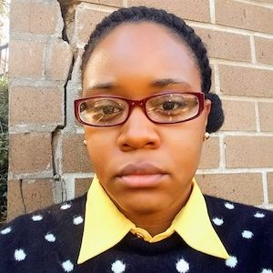 headshot of Menika weaing glasses standing against a brick wall in a blue and white polka dot sweater and yellow collar shirt