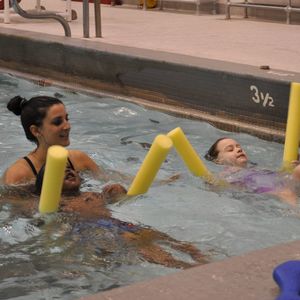 A swim instructor oversees two children in the pool with flotation noodles.