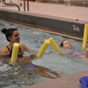 Two kids swimming with noodles as a swim instructor watches them.