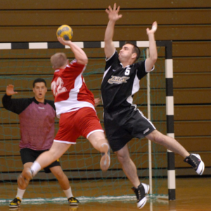 A player jumps into the air to shoot while the goalie and defender try to get in the way.