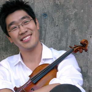 headshot of a smiling man with black hair, glasses, and a white polo shirt, leaning against a concrete wall and holding a violin