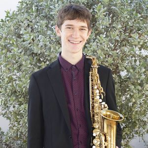 smiling man holding a saxophone and wearing a suit, standing in front of a bush