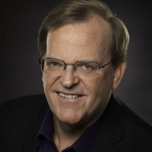 headshot of a man with dark blond hair, glasses, and wearing a purple shirt and black blazer