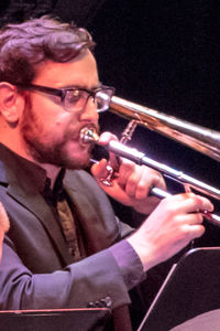 headshot of a man with glasses and beard playing a trombone