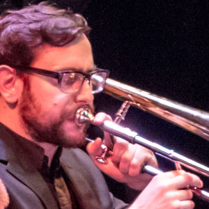 headshot of man with glasses and beard playing a trombone