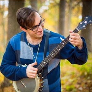 Man with glasses and a blue patchwork sweater smiling and playing a banjo in a forest