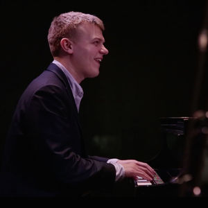 Blond man in a black suit smiling and playing the piano