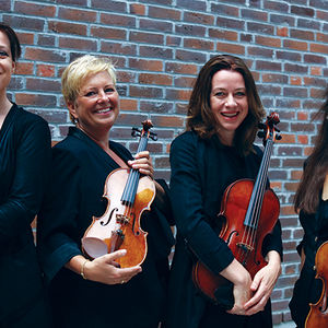 4 women with string instruments dressed in black, standing in front of a brick wall