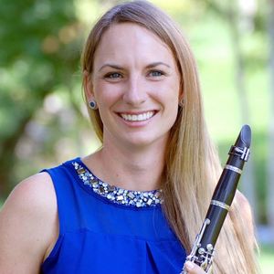 headshot of smiling blonde woman with a blue shirt and blue stone earrings, holding a clarinet