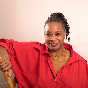 woman with small dreadlocks, a red button-down shirt, and a gold beaded necklace sitting with her right arm over the back of a wooden chair
