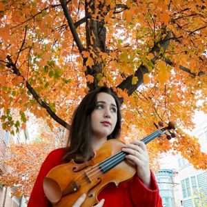 woman with brown hair and a red shirt holding a violin and standing in front of a tree with autumn foliage