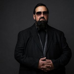 man with a long black beard, a black suit, and black aviator sunglasses standing with his hands folded